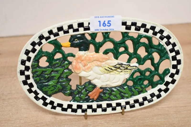A sweet vintage cast iron duck trivet, of oval form featuring a hand painted duck against a green foliage background with a black and white checkered boarder