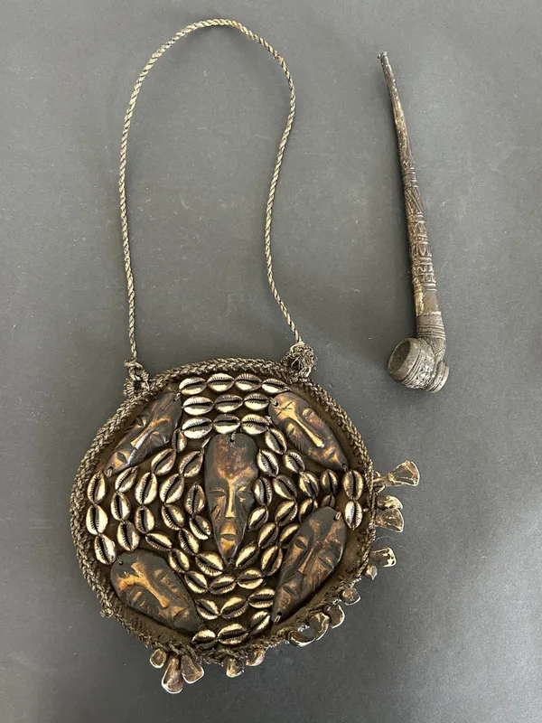 A vintage African ceremonial or tribal pendant (chest plate) with carved faces and cowrie shell decoration etc (with wear) together with an antique smoking pipe