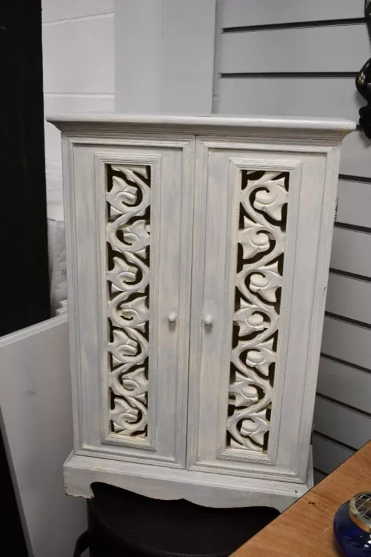 A vintage wooden cream-coloured storage cabinet, featuring two doors with carved foliate detailing, having four internal shelves, unmarked,